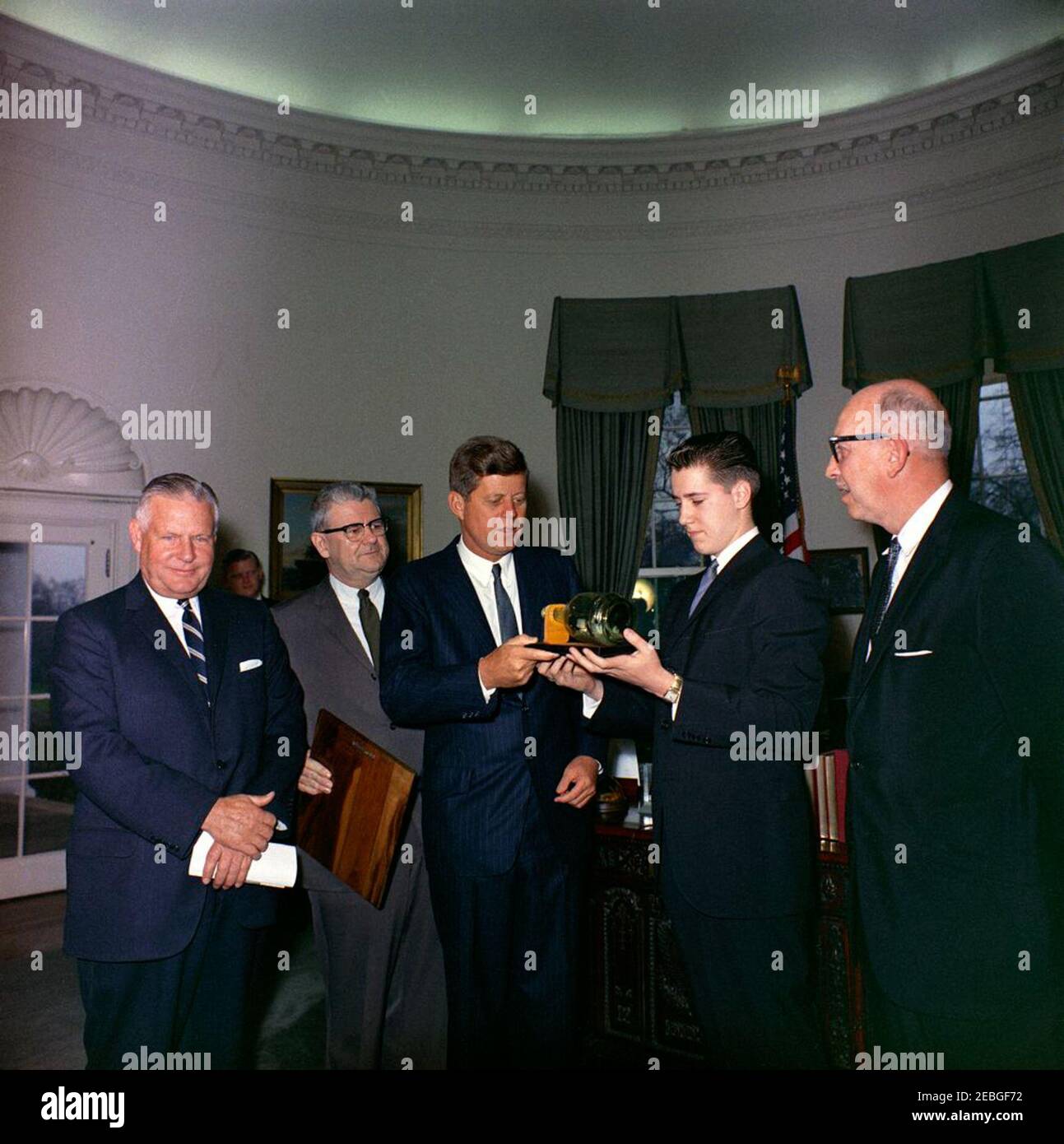 Visit of the Boysu0027 Clubsu0027 u0022Boy of the Year,u0022 Stephen Lutz, 10:57AM. President John F. Kennedy (center) holds a scale model, enclosed inside a bottle, of PT Boat 109, presented to him by the Boysu0027 Clubs of Americau0027s (BCA) u0022Boy of the Yearu0022 Stephen Lutz of Newark, New Jersey. (L-R) Albert L. Cole, President of the BCA; unidentified man (in back, partially hidden); John M. Gleason, National Director of the BCA; President Kennedy; Stephen Lutz; and Charles R. Messier, Executive Director of the Boysu0027 Clubs of Newark, New Jersey. Oval Office, White House, Stock Photo