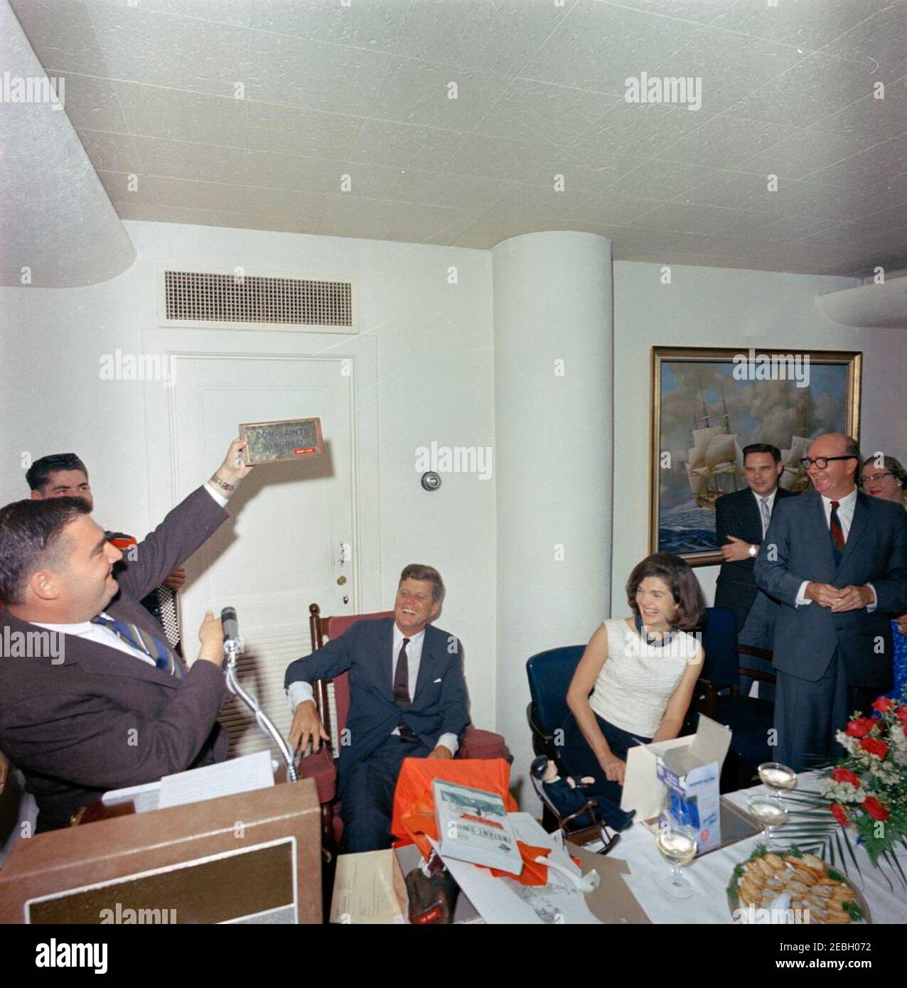 Surprise birthday party for President Kennedy, given by the White House staff, 5:45PM. Press Secretary Pierre Salinger (far left) holds up a plaque that reads u201cComplaints Ignored / Henry Luce,u201d during a surprise party thrown by members of the White House staff in honor of President John F. Kennedyu2019s 46th birthday. President Kennedy (sitting in rocking chair) and First Lady Jacqueline Kennedy laugh as Salinger opens gifts. Presidential Aide Dave Powers stands second from right (wearing glasses); others are unidentified. Navy Mess Hall, White House, Washington, D.C. Stock Photo