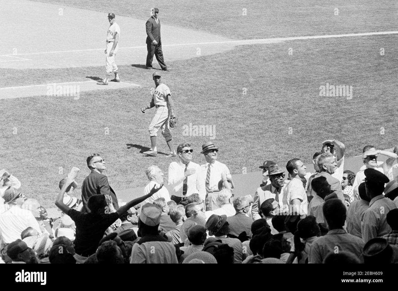 1962 All Star Baseball Game at D.C. Stadium, 12:52PM. President John F. Kennedy (wearing sunglasses) and others watch the 32nd Major League Baseball (MLB) All-Star Game at D.C. Stadium, Washington, D.C. First baseman for the National League, Ernie Banks (Chicago Cubs), and first base coach for the American League, James Barton u0022Mickeyu0022 Vernon (manager of the Washington Senators), stand on the field in background. Also pictured (in the stands): Commissioner of Baseball, Ford C. Frick; Special Assistant to the President, Dave Powers; Associate Press Secretary, Andrew T. Hatcher; member Stock Photo