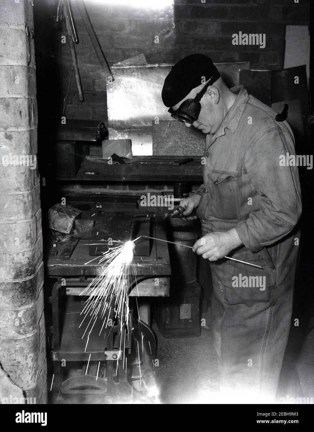 1950s, historical, an artisan workman in overalls and wearing goggles and a berry on his hat in a small workshop doing metalwork or welding. Welding is the process of joining metals together. Fusion is the generic sheet metal fabrication term for joining together metals of similar compositions and melting points. A pool of molten material called the weld pool is formed due to the high melting points of the workpieces. Stock Photo