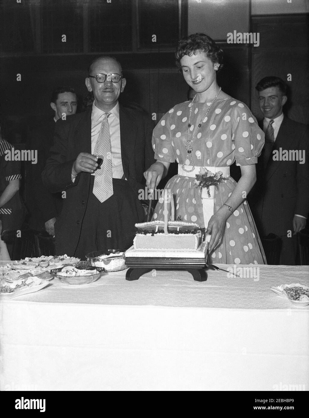 1950s, historical, standing beside her proud grandfather, an elegant young woman wearing a stylish polka dot or spotted dress cutting her birthday cake at her birthday party in a function room of a hotel, England, UK. Stock Photo