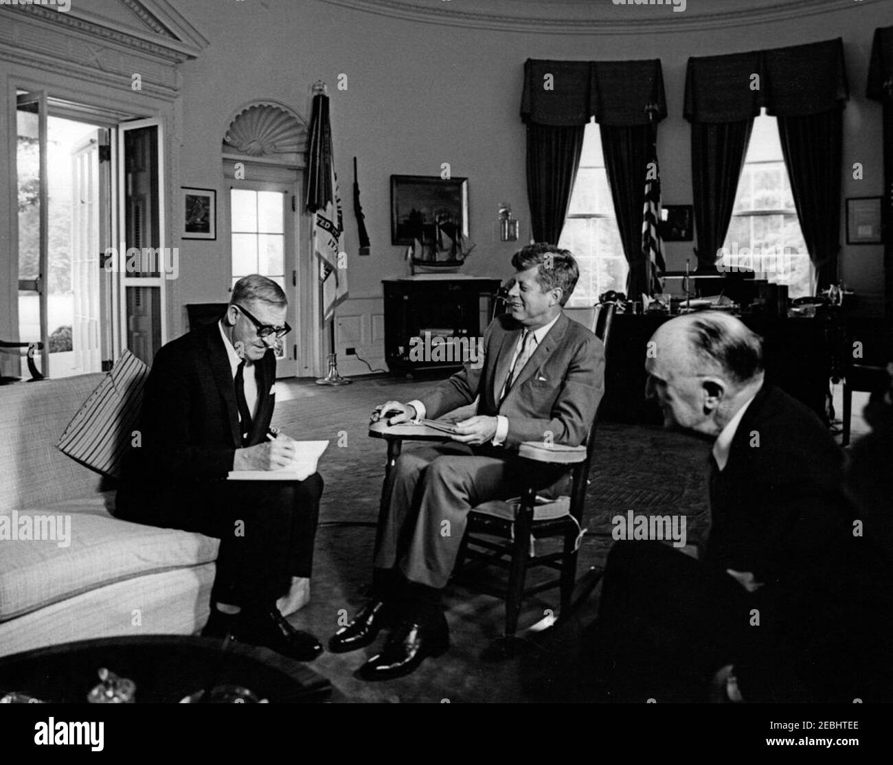 Meeting with Arthur A. Calwell, Leader of the Labor Party of Australia, 12:05PM. President John F. Kennedy (in rocking chair, holding a pen in one hand and a book in the other) meets with Leader of the Australian Labor Party, Arthur A. Calwell (left, writing in a book); Ambassador of Australia, Sir Howard Beale, sits at right. Oval Office, White House, Washington, D.C Stock Photo