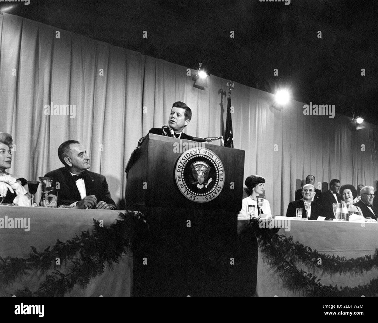 National Cultural Center Fund-raising benefit, 9:23PM. President John F. Kennedy (at lectern) delivers remarks at a fundraising dinner and closed-circuit telecast of u0022An American Pageant of the Artsu0022 for the National Cultural Center. Seated (L-R): chairman of the dinner committee for the National Cultural Center, Suzanne Gardner; Vice President Lyndon B. Johnson; First Lady Jacqueline Kennedy; Chairman of the Board of Trustees for the National Cultural Center, Roger L. Stevens; Lady Bird Johnson; and Chief Justice of the Supreme Court, Earl Warren. National Guard Armory, Washington, Stock Photo