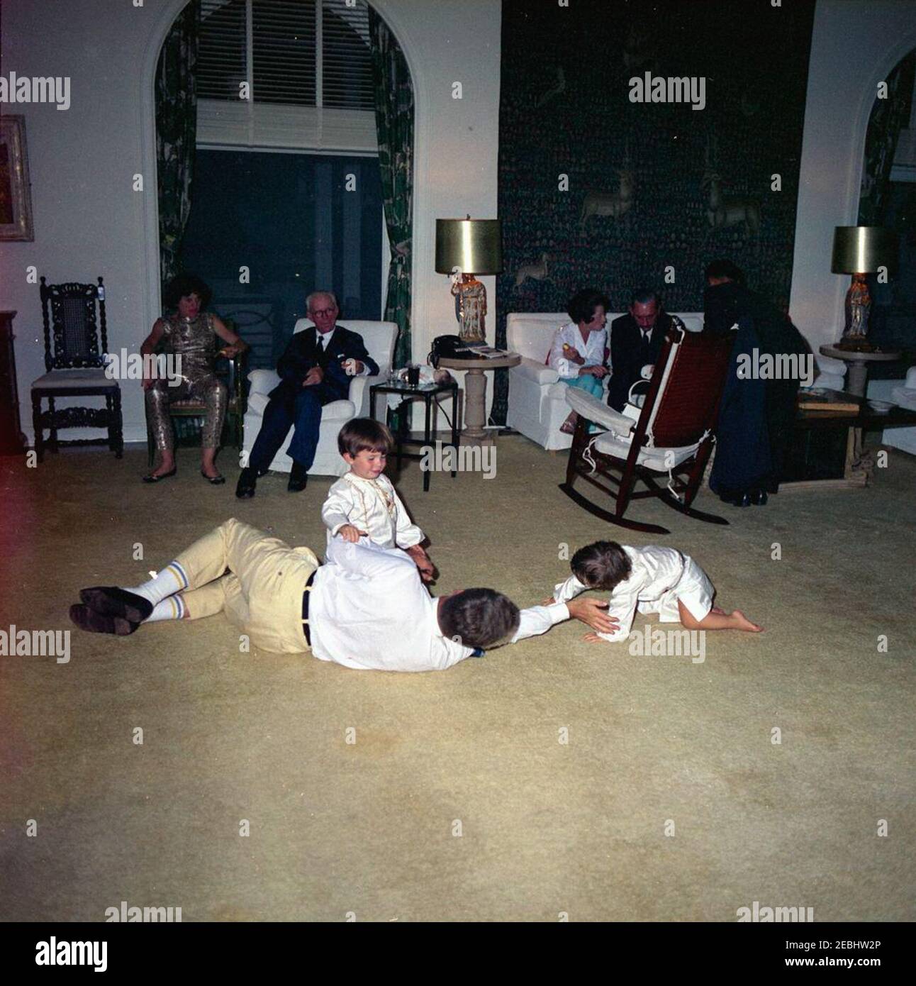 Christmas Day, Palm Beach. President John F. Kennedy (lying on floor) plays with his son, John F. Kennedy, Jr. (right), and nephew, Anthony Radziwill (left), on Christmas. Seated in background (L-R): Eunice Kennedy Shriver, Joseph P. Kennedy, Sr., Rose Fitzgerald Kennedy, and Prince Stanislaus Radziwill of Poland. Residence of C. Michael Paul, Palm Beach, Florida. Stock Photo