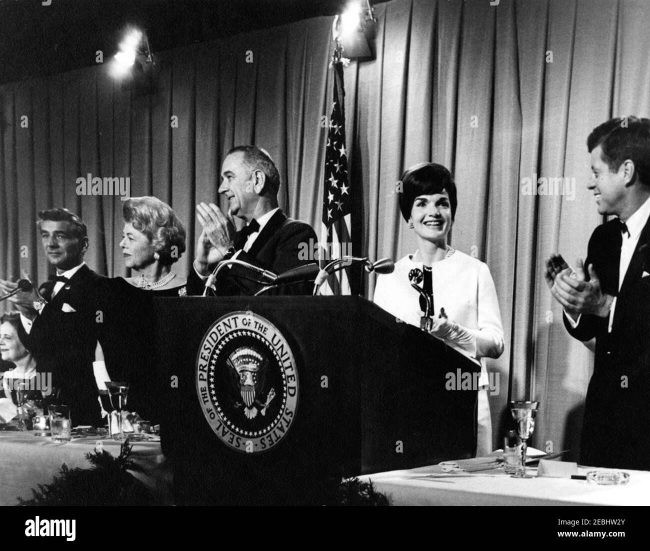 National Cultural Center Fund-raising benefit, 9:23PM. President John F. Kennedy, First Lady Jacqueline Kennedy, and others attend a fundraising dinner and closed-circuit telecast of u0022An American Pageant of the Artsu0022 for the National Cultural Center. Left to right: Nina Warren (seated), wife of Earl Warren, Chief Justice of the Supreme Court; master of ceremonies, composer and conductor Leonard Bernstein; chairman of the dinner committee for the National Cultural Center, Suzanne Gardner; Vice President Lyndon B. Johnson; Mrs. Kennedy; and President Kennedy. National Guard Armory, Was Stock Photo