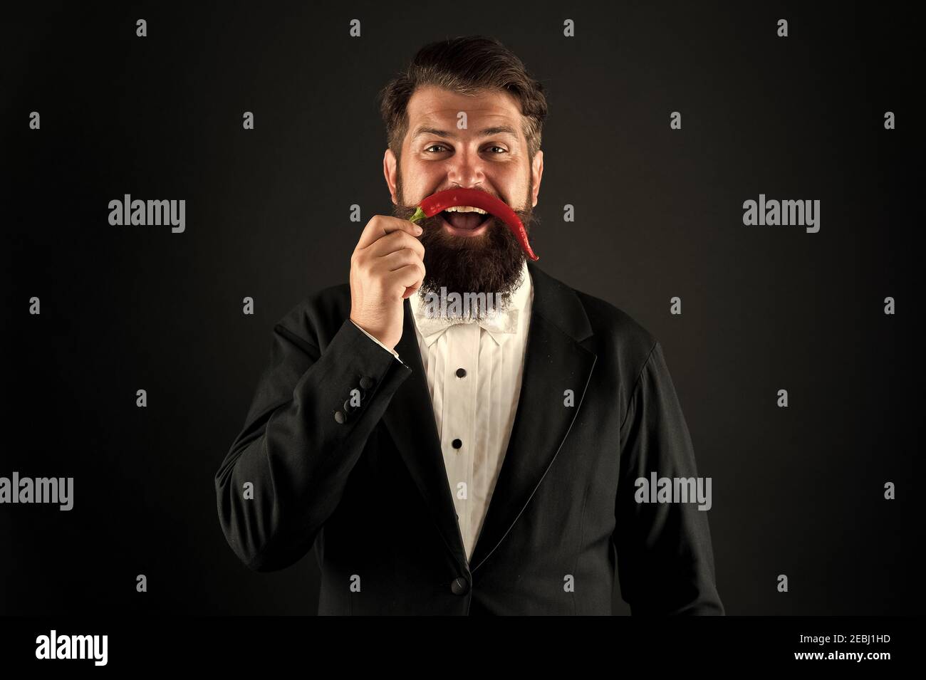 Handsome macho likes spicy taste. Man hold pepper harvest. Barber funny face. Pepper mustache. Bearded businessman hold chilli red pepper in hand. Guy hold Hot chilli pepper black background. Stock Photo
