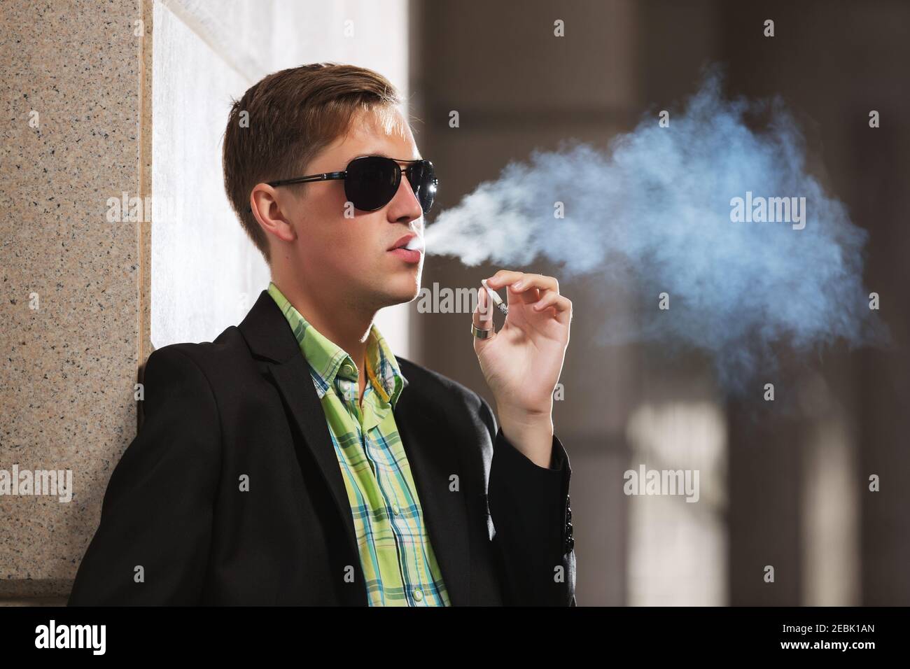Young man in black blazer and sunglasses smoking a cigarette on city street Stock Photo
