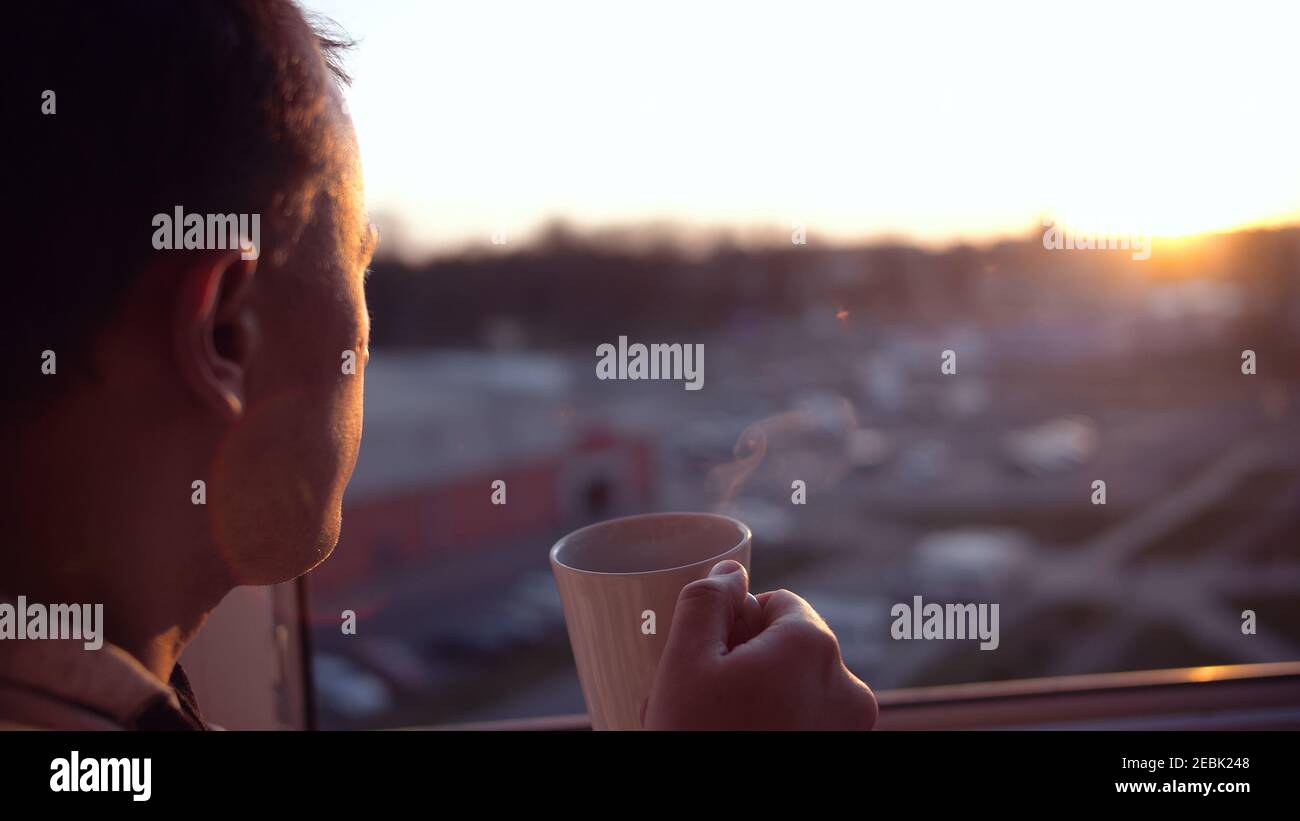 A man stands at the window and looks at the sunset, drinks tea from a mug, close-up Stock Photo