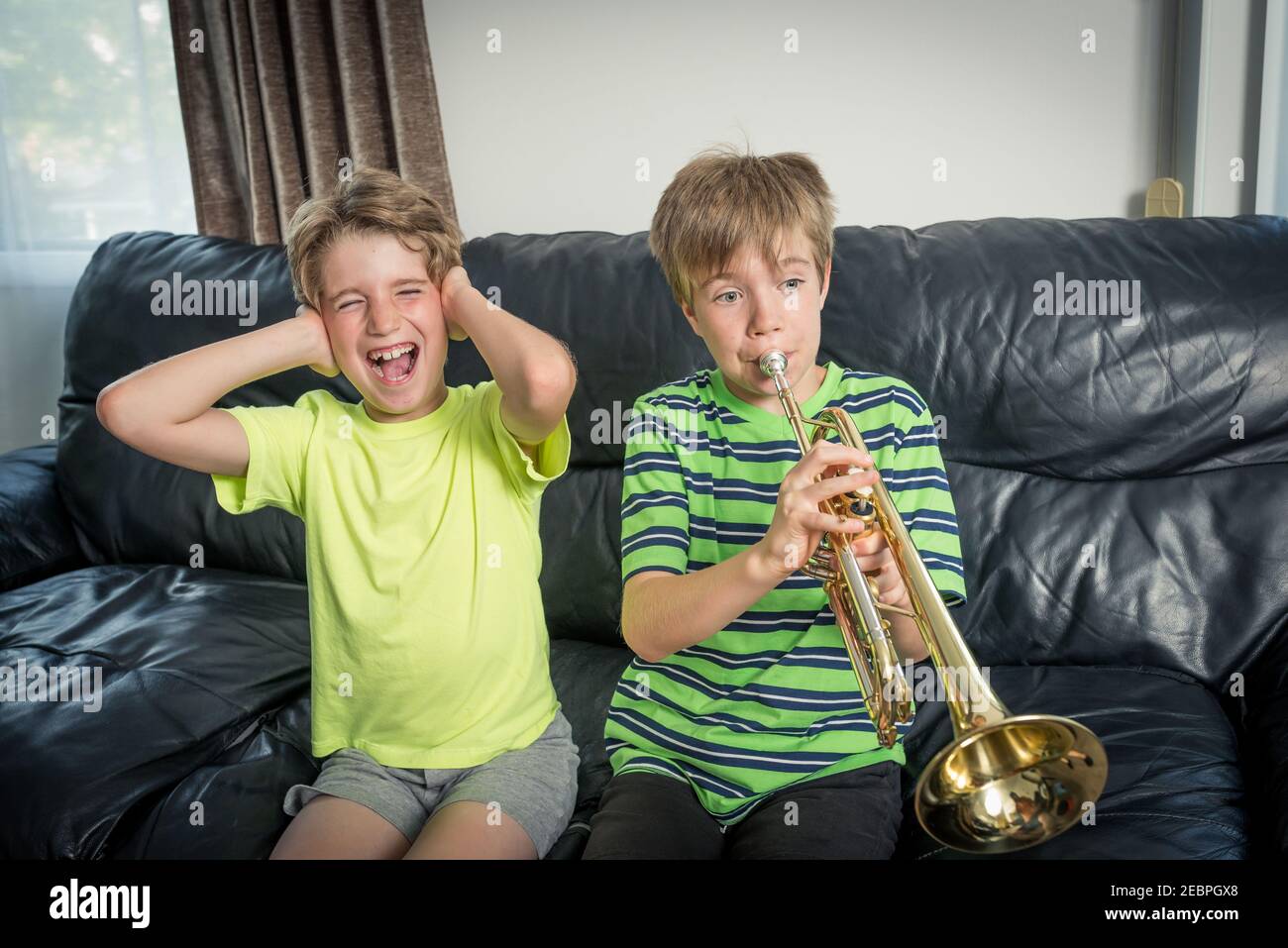Two kids sitting on a sofa. One is playing a trumpet and the other child annoyed is covering his ears with his hands Stock Photo