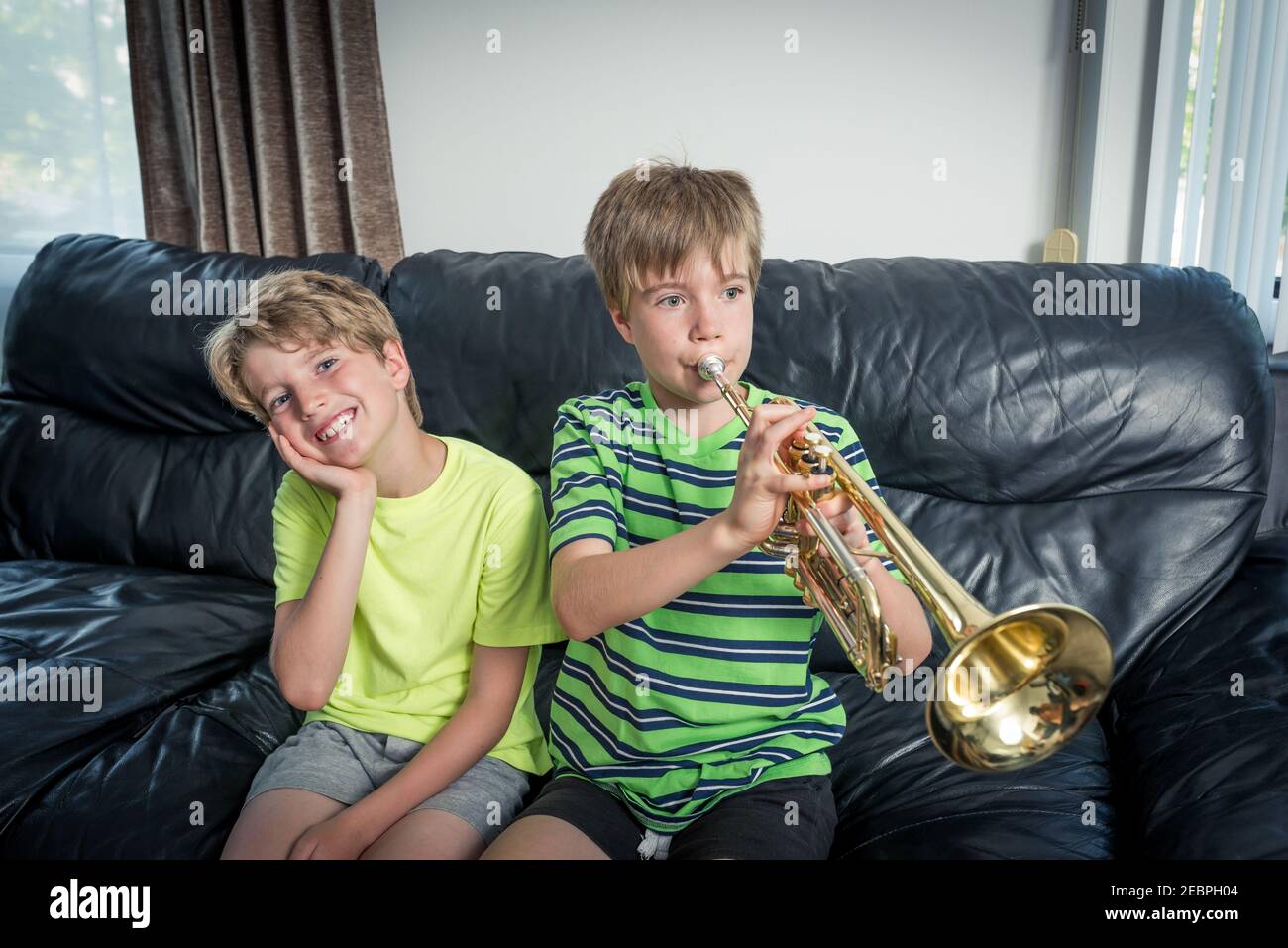 Two kids sitting on a sofa.  One is playing a trumpet and the other child is listening and smiling. Stock Photo