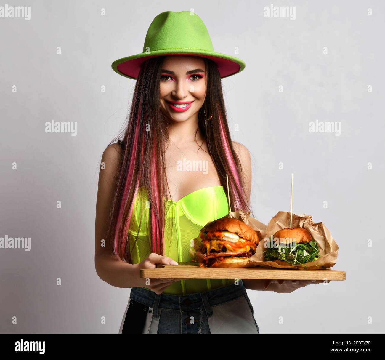 Young woman in jeans, corset and wide-brimmed hat stands holding two craft big burger sandwiches on wooden tray Stock Photo