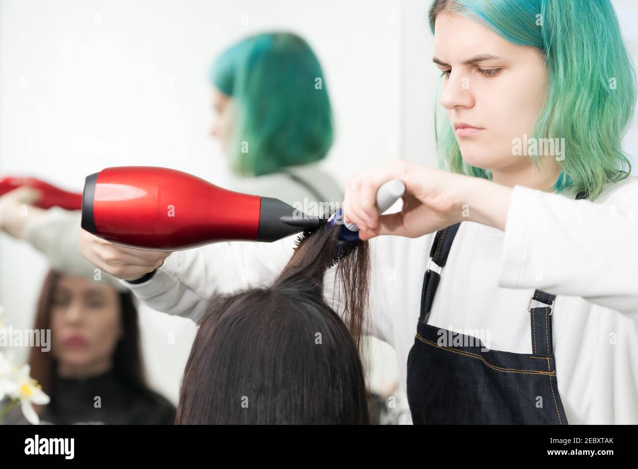 Professional hairdresser with green hair dries hair to client with red hairdryer and blue comb in beauty salon. Stock Photo