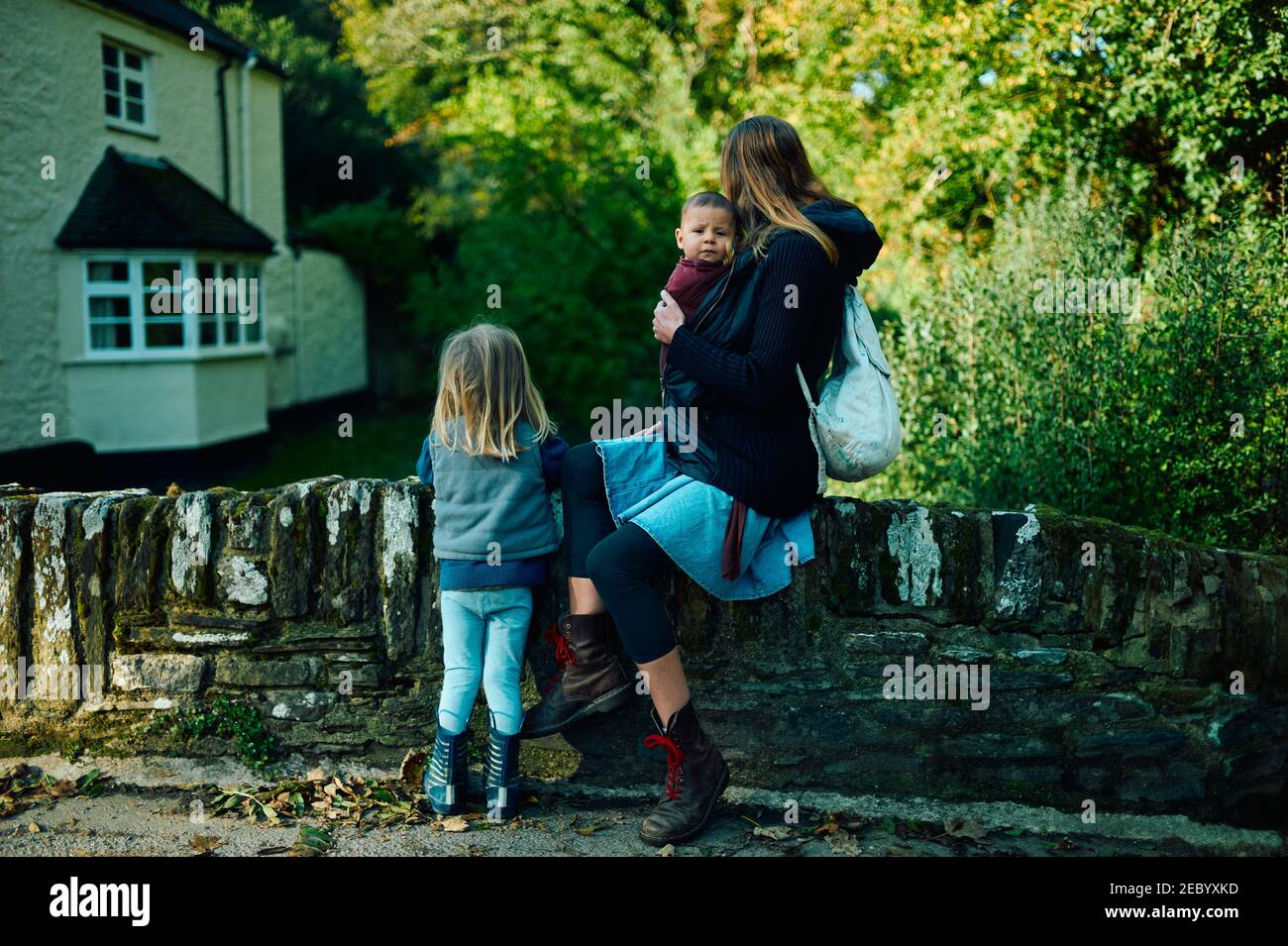 A young mother is sitting on a wall in the countryside with her preschooler and her baby in a sling Stock Photo