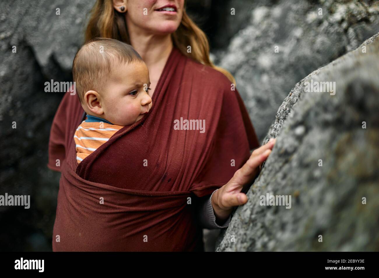 A young mother with her baby in a sling is walking by rocks outdoors in nature Stock Photo