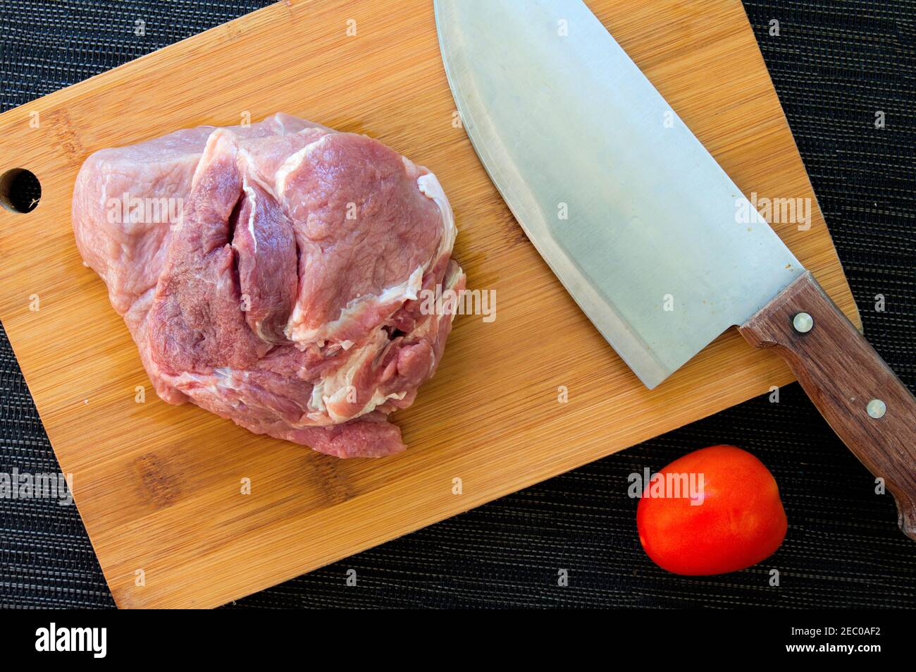Meat and tomato on wooden board. Cutting meat by chef knife. Beef or pork for cooking. Kitchen table top view with meat and vegetables. Stainless stee Stock Photo