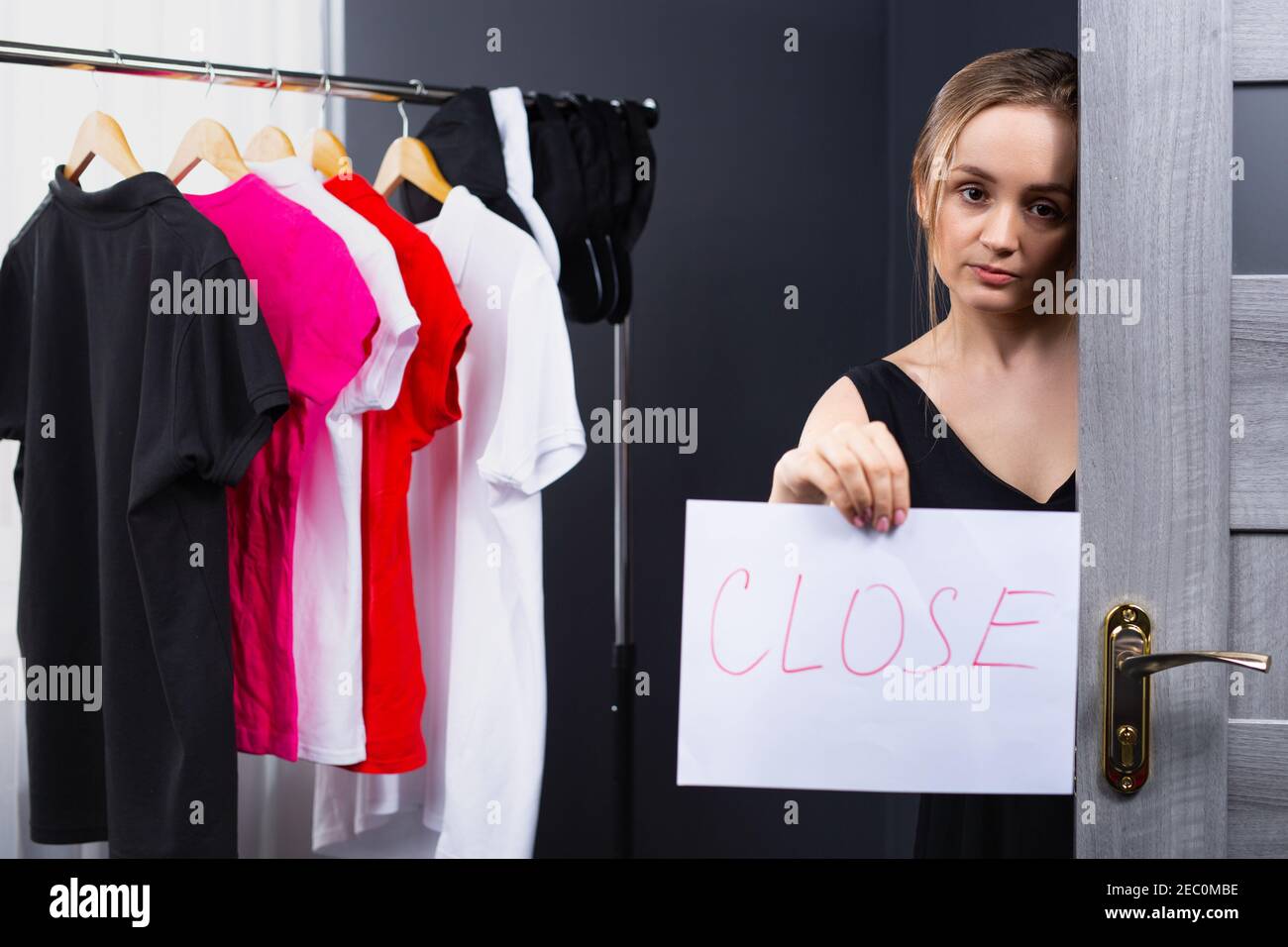 A kind and beautiful blonde, sad, woman working as a consultant in a clothing store indicates that the store is closed on a signboard. Photo indoor Stock Photo