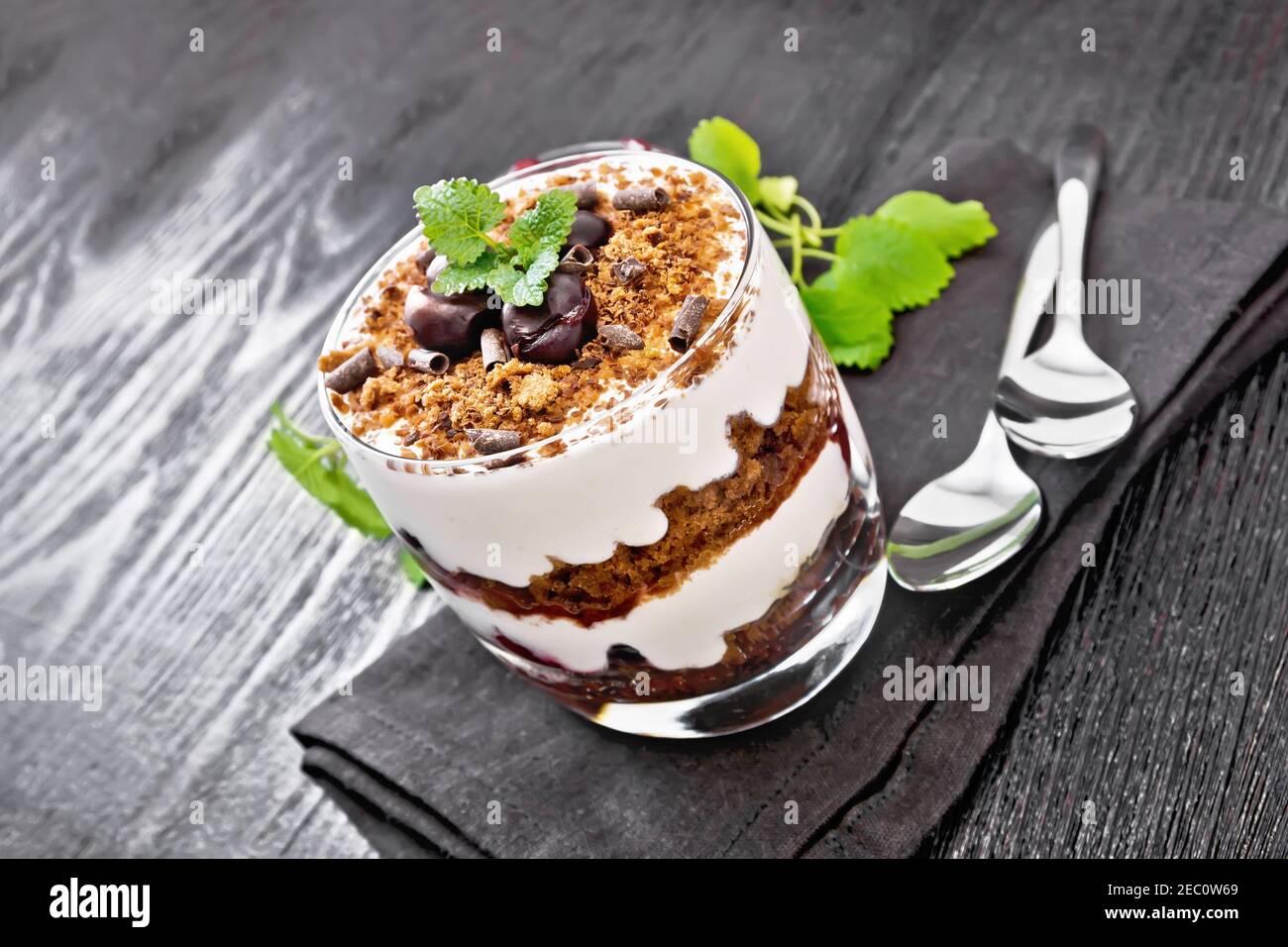 Dessert Black Forest of cherries, chocolate biscuit and soft cottage cheese with cream in a glass on napkin, mint, berries in a bowl and a spoon on da Stock Photo