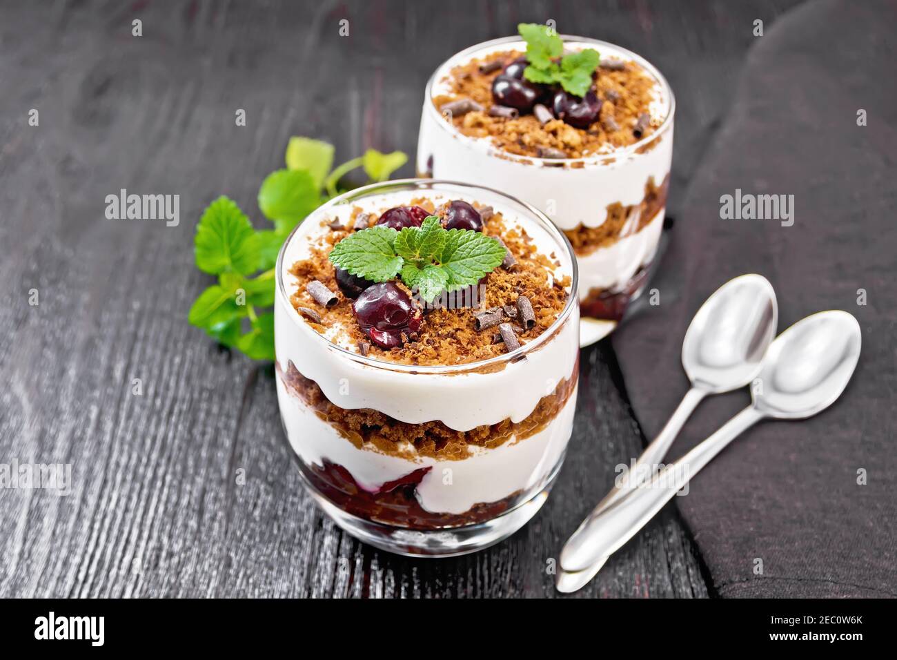 Dessert Black Forest of cherries, chocolate biscuit and soft cottage cheese with cream in two glasses, a towel, mint and a spoon on dark wooden board Stock Photo