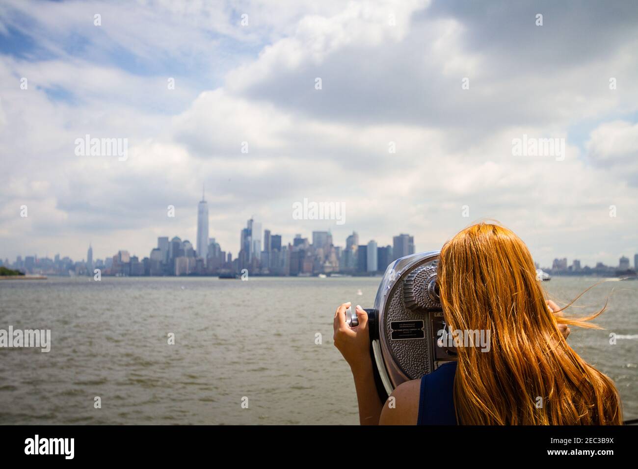 A young woman with red hair is looking at the cityscape of New York City through a binocular on Liberty Island Stock Photo