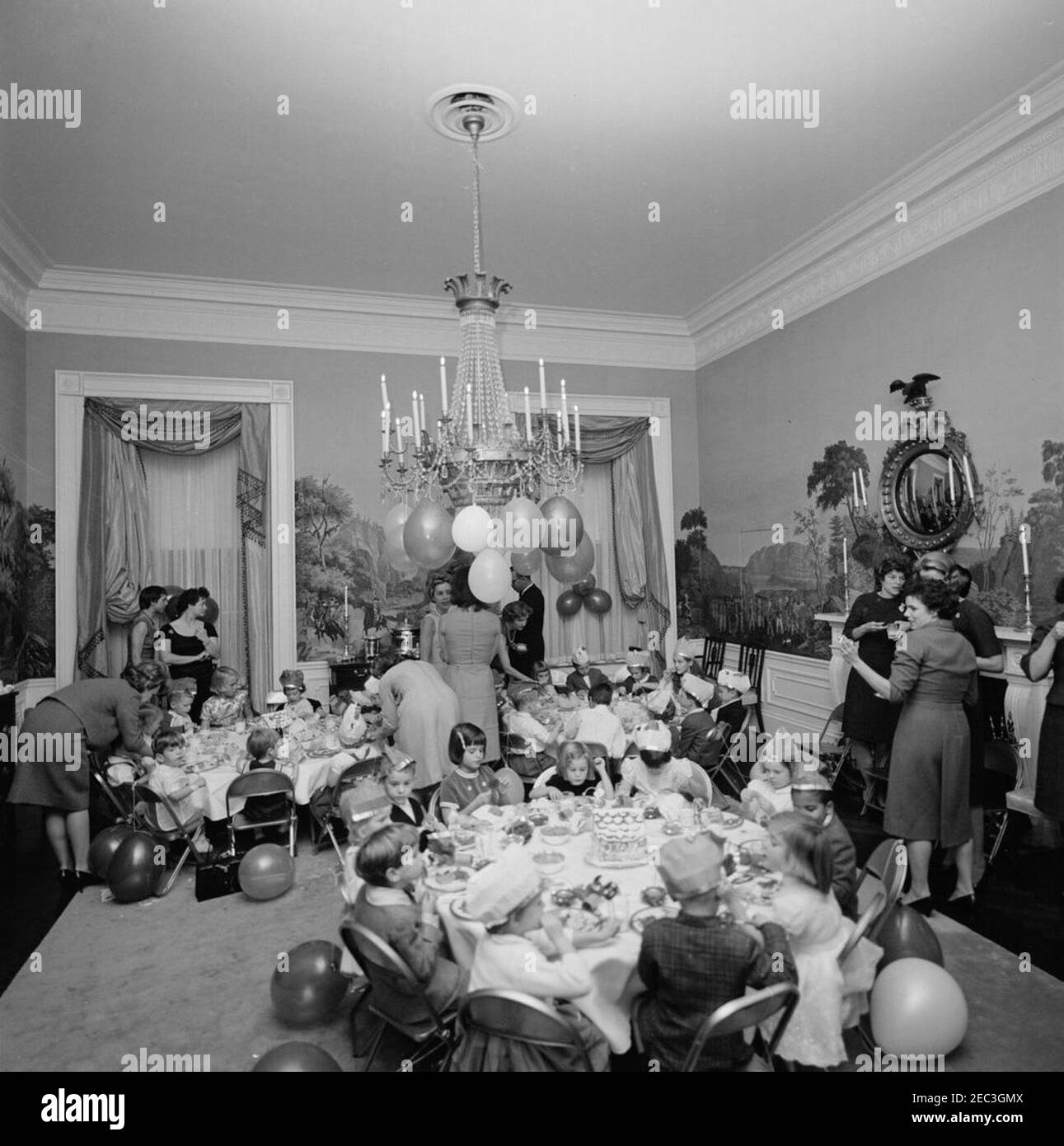 Birthday party for Caroline Kennedy and John F. Kennedy, Jr.. Young children sit at tables in the Presidentu2019s Dining Room (Residence) of the White House, Washington, D.C., during a joint birthday party for Caroline Kennedy and John F. Kennedy, Jr.; First Lady Jacqueline Kennedy (back to camera) stands at center in background. Caroline (right) sits at table in foreground with Maria Shriver (seated in center, holding lollipop) and Avery Hatcher, son of Associate Press Secretary, Andrew T. Hatcher (seated right of Caroline, partially hidden); John, Jr. (left, facing the camera), sits at tabl Stock Photo