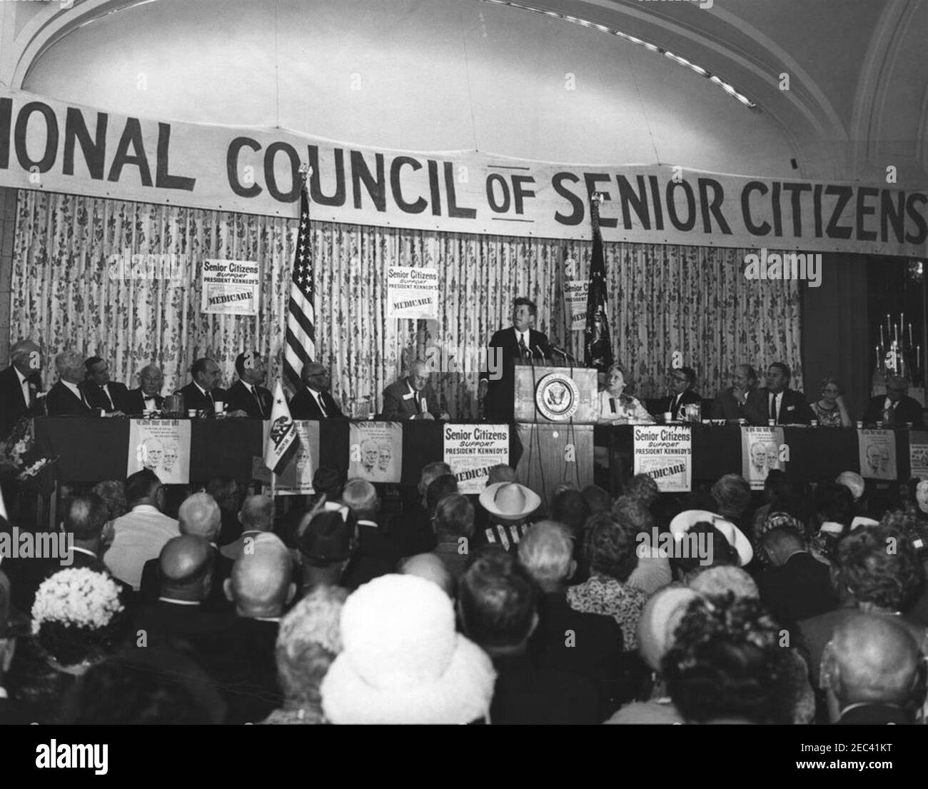 Address to the National Convention of the National Council of Senior Citizens, 11:47AM. President John F. Kennedy (at lectern) delivers an address to attendees of the National Convention of the National Council of Senior Citizens at the Willard Hotel, Washington D.C. On stage (L-R): Council Vice President, Burt Garnett; three unidentified; Secretary of the Department of Health, Education, and Welfare, Anthony J. Celebrezze; Senator Birch Evans Bayh, Jr. of Indiana; Director of Special Projects for the Council, Lawrence A. Oxley; Council Chairman, John Fitzpatrick; President Kennedy; two uniden Stock Photo