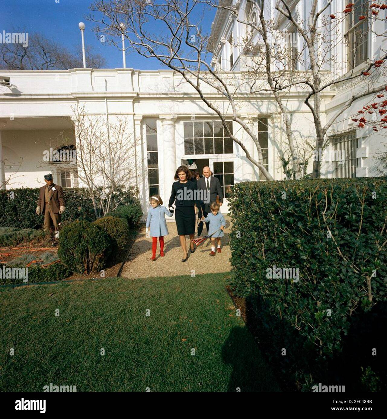 Jacqueline Kennedy (JBK) departs White House. Former first lady, Jacqueline Kennedy, and her children, Caroline Kennedy and John F. Kennedy, Jr., depart the White House for the last time. Special Assistant to President John F. Kennedy, Dave Powers, follows behind; Princess Lee Radziwill of Poland (sister of Mrs. Kennedy) walks through the Palm Room door. Rose Garden, White House, Washington, D.C. Stock Photo