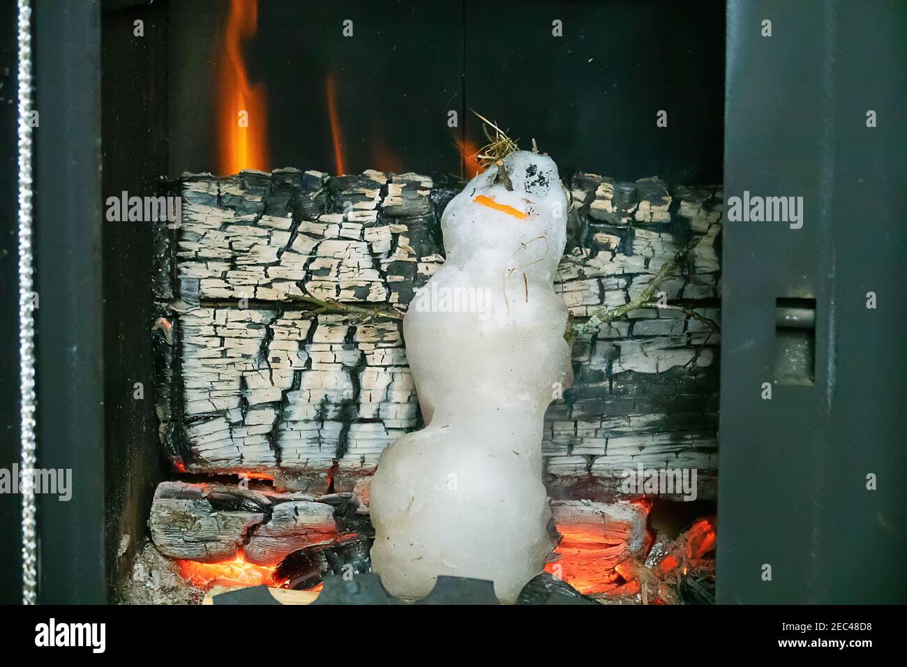 Little snowman melts quickly in hot stove against background of firewood. Concept of victory of heat over cold (spring over winter), heat-loving human Stock Photo