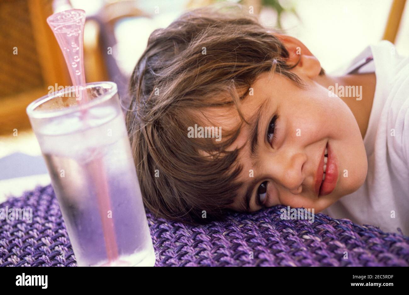 A small boy resting his head on the table, cheekily looking at the camera at an outdoor hotel restaurant in Crete, Greece. Stock Photo