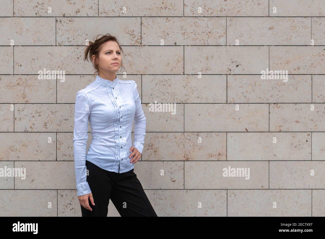 Attractive woman in white shirt standing on marble brick wall background and looking ahead. Successful and purposeful woman concept. Copy space. Stock Photo