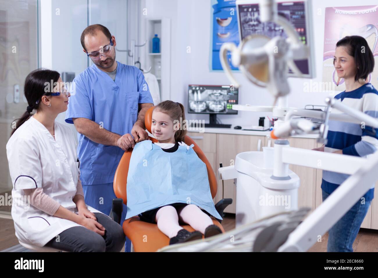 Little girl wearing dental bib sitting on chair in dentist office during tooth examining .Child with her mother during teeth check up with stomatolog sitting on chair. Stock Photo