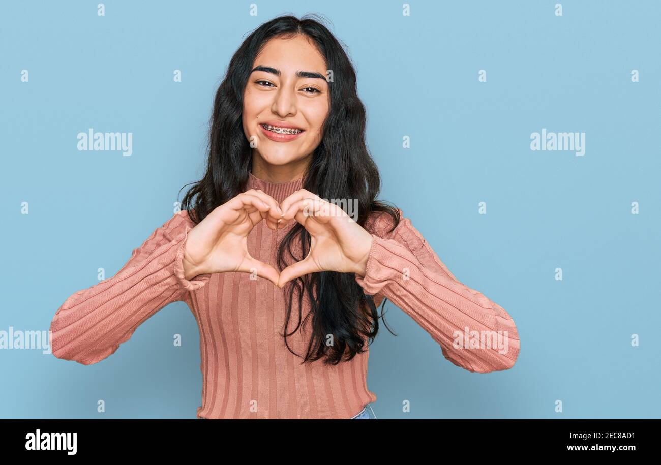 Hispanic teenager girl with dental braces wearing casual clothes smiling in love doing heart symbol shape with hands. romantic concept. Stock Photo