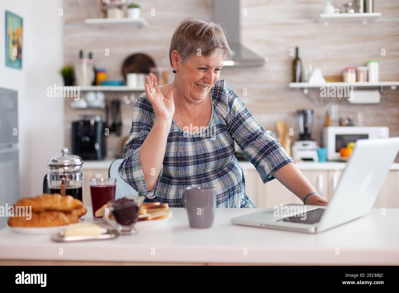 Retired woman waving during online meeting with family on video call using laptop in kitchen having breakfast. Elderly person using internet online chat tech, pc webcam for virtual conference call Stock Photo