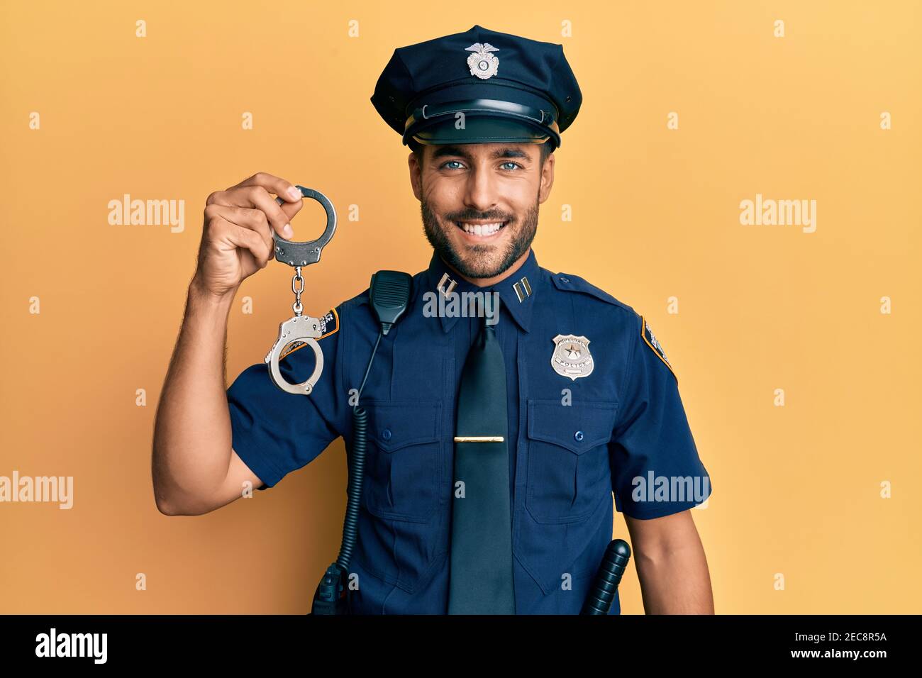 Handsome hispanic man wearing police uniform holding metal handcuffs looking positive and happy standing and smiling with a confident smile showing te Stock Photo