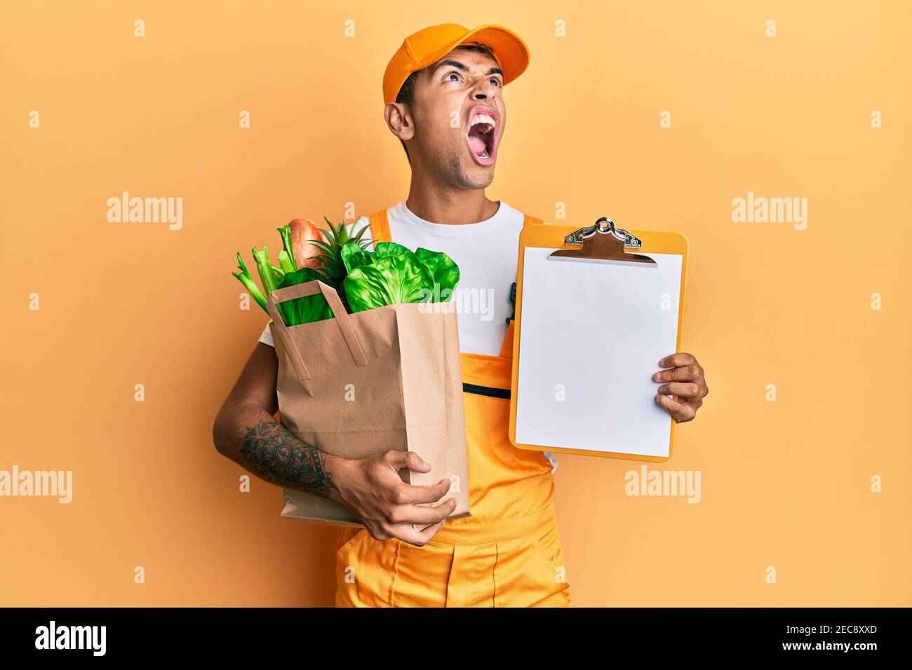 Young handsome african american man wearing courier uniform with groceries from supermarket and clipboard angry and mad screaming frustrated and furio Stock Photo