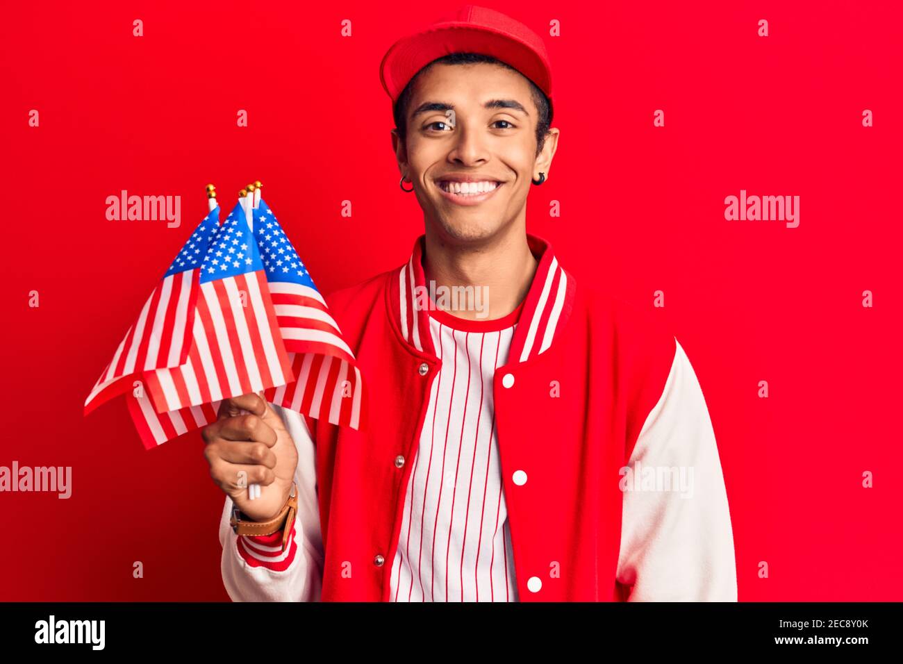 Young african amercian man wearing baseball uniform holding america flags looking positive and happy standing and smiling with a confident smile showi Stock Photo