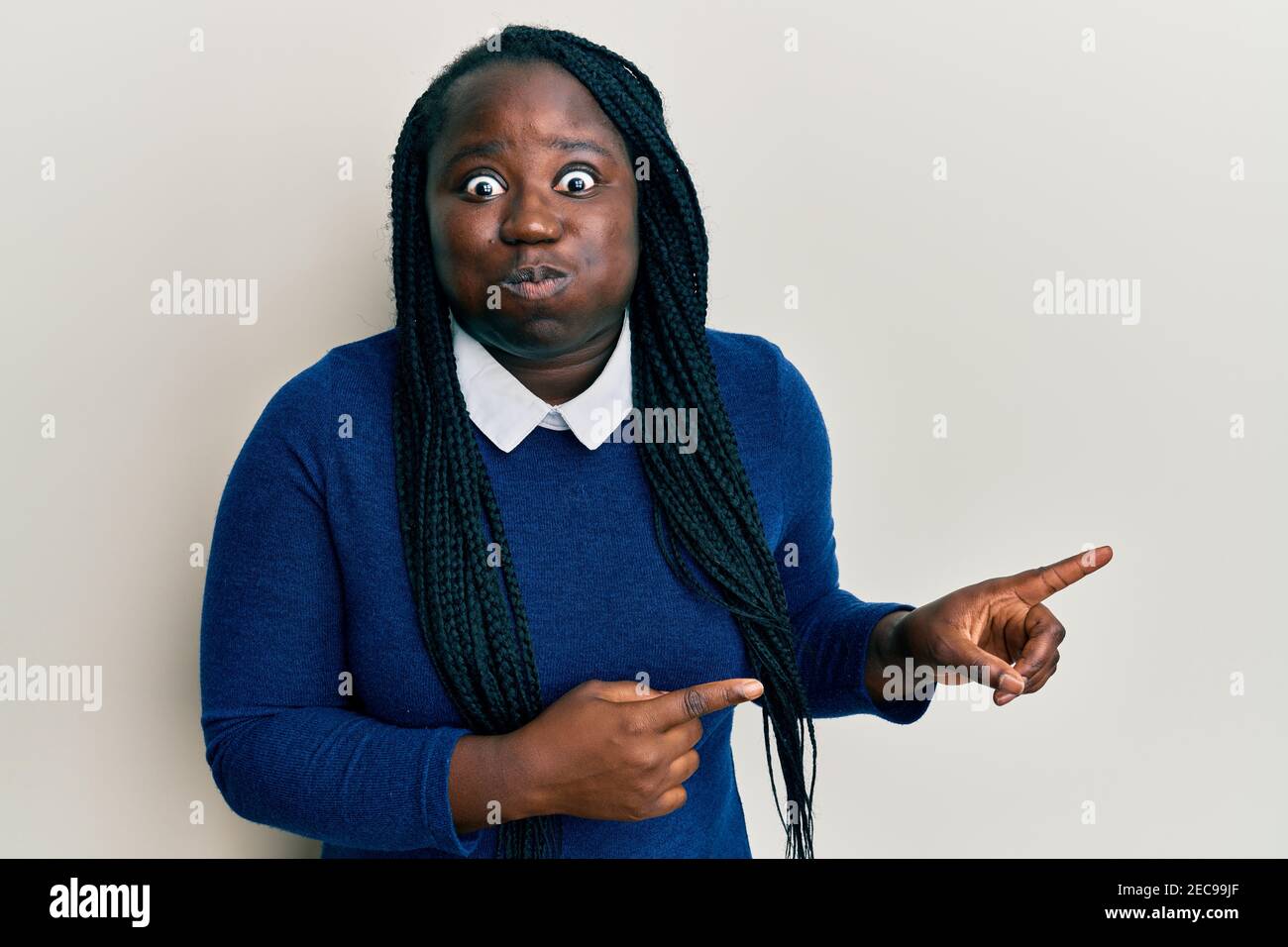 Young black woman with braids pointing up with fingers to the side puffing cheeks with funny face. mouth inflated with air, catching air. Stock Photo