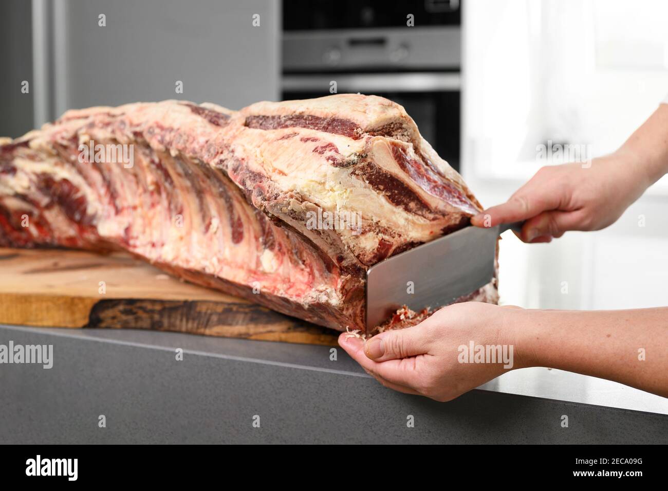 Chef is preparing prime rib steaks from a large prime beef rib roast with multiple bones. The meat is on a wooden board which is sitting on a modern kitchen counter. High quality photo Stock Photo