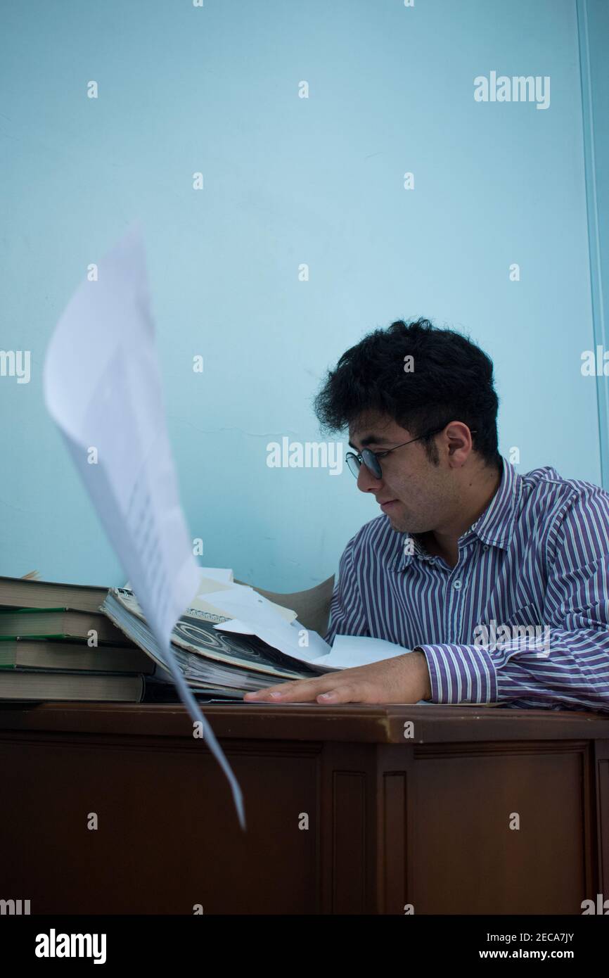 Young man wearing striped shirt working in the office with many books and sheets on a wooden desk with a blue background Stock Photo