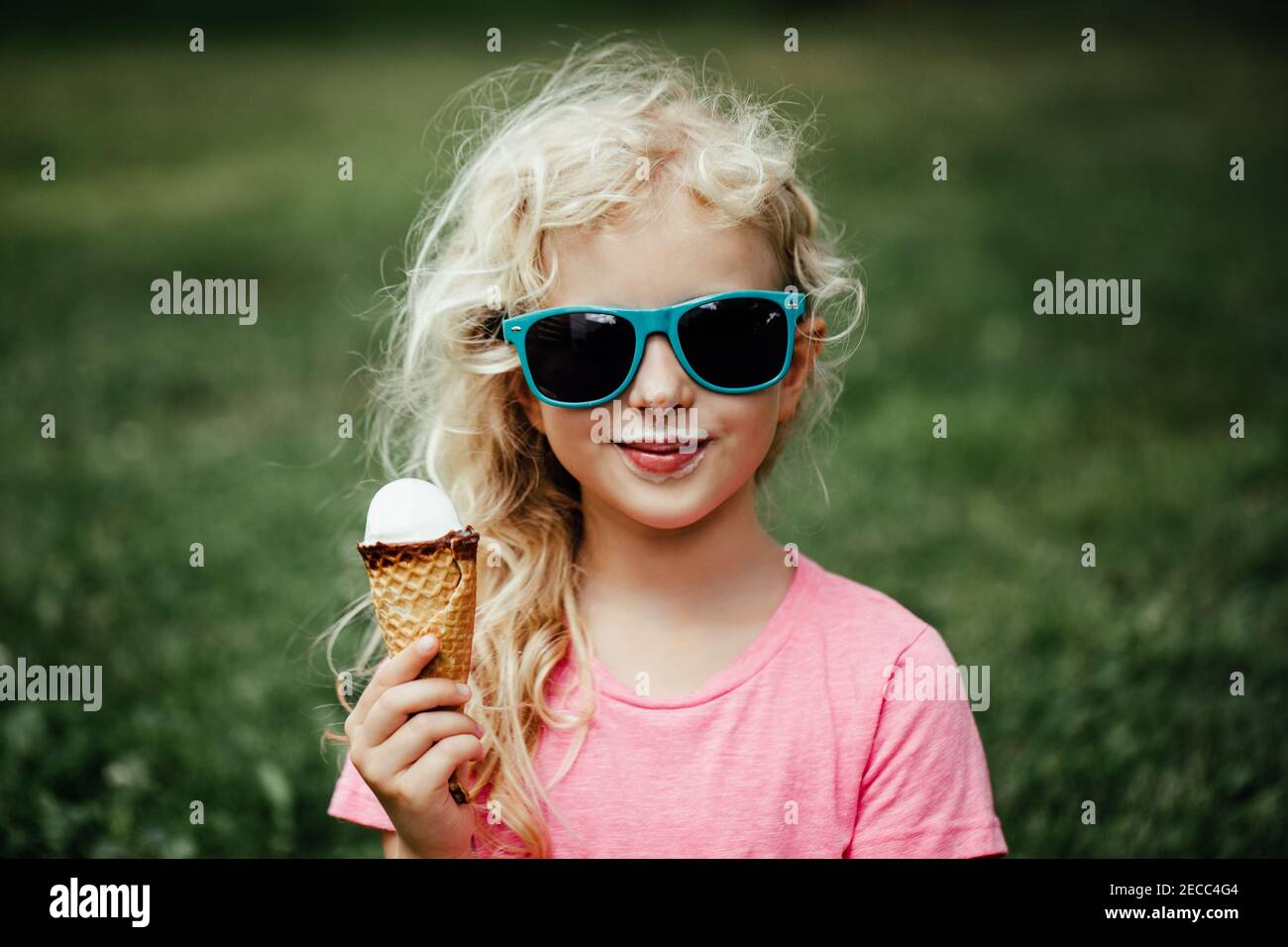 Cute funny adorable girl in sunglasses with dirty nose and moustaches eating ice cream from waffle cone. Happy cool hipster child eating tasty sweet c Stock Photo