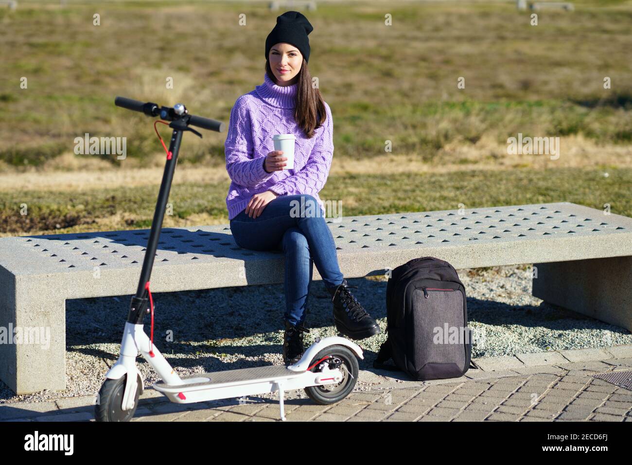 Female student with electric scooter taking a coffee break sitting on a bench outside her college. Stock Photo