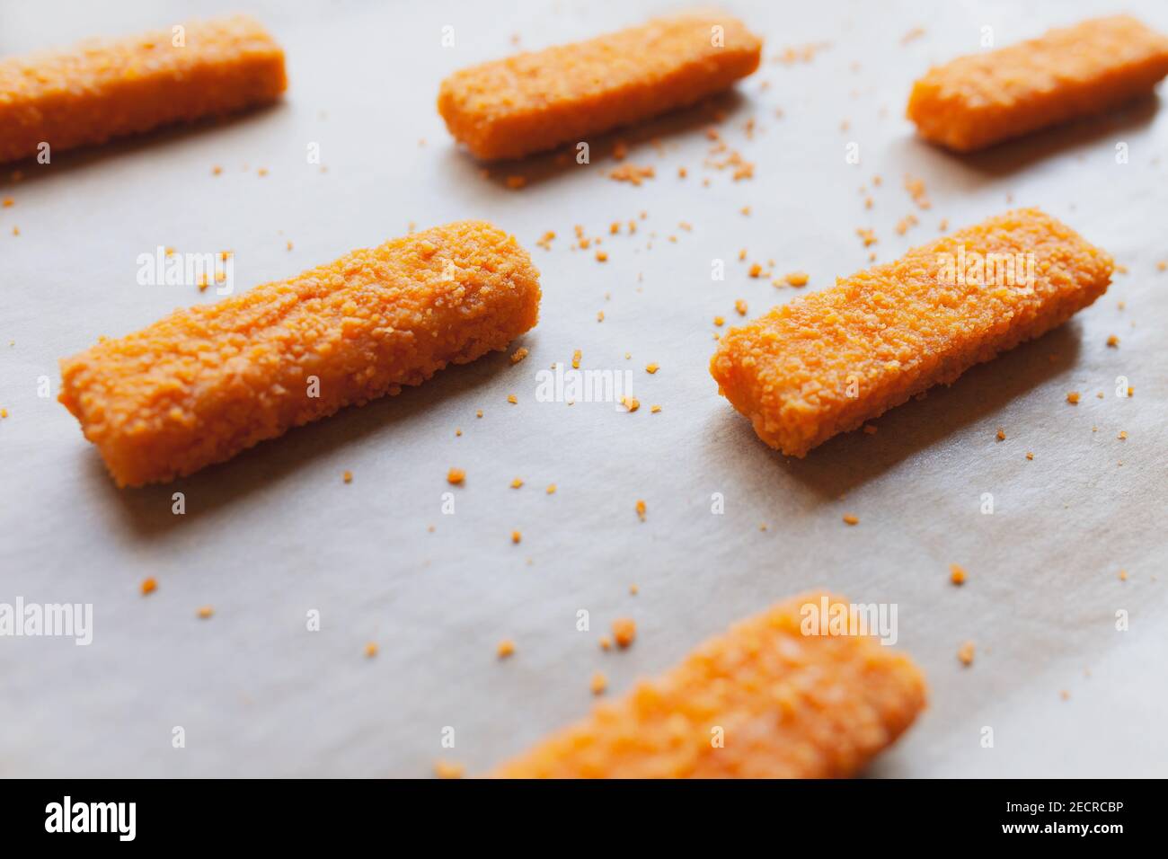 Raw frozen fish fingers on parchment paper, ready for baking, with some crumbs Stock Photo