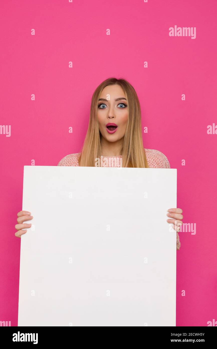 Surprised young woman is holding white big empty banner. Front view. Waist up studio shot on pink background. Stock Photo