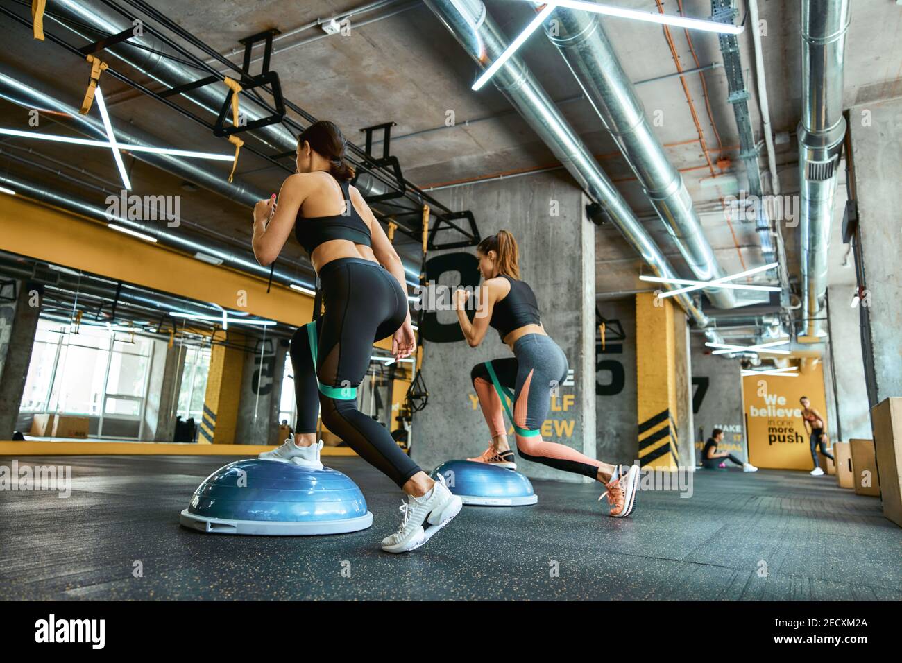 Full length of two young athletic women in sportswear exercising together  with resistance fitness bands at crossfit gym, doing squats and training  legs. Workout, sport, wellness and healthy lifestyle Stock Photo -