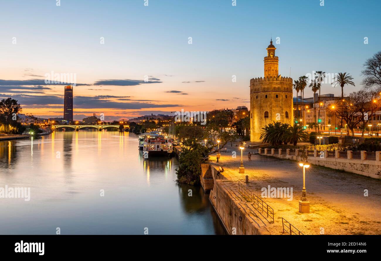 Boardwalk at the river Rio Guadalquivir with illuminated Torre del Oro, in the back Torre Sevilla, sunset, blue hour, Sevilla, Andalusia, Spain Stock Photo