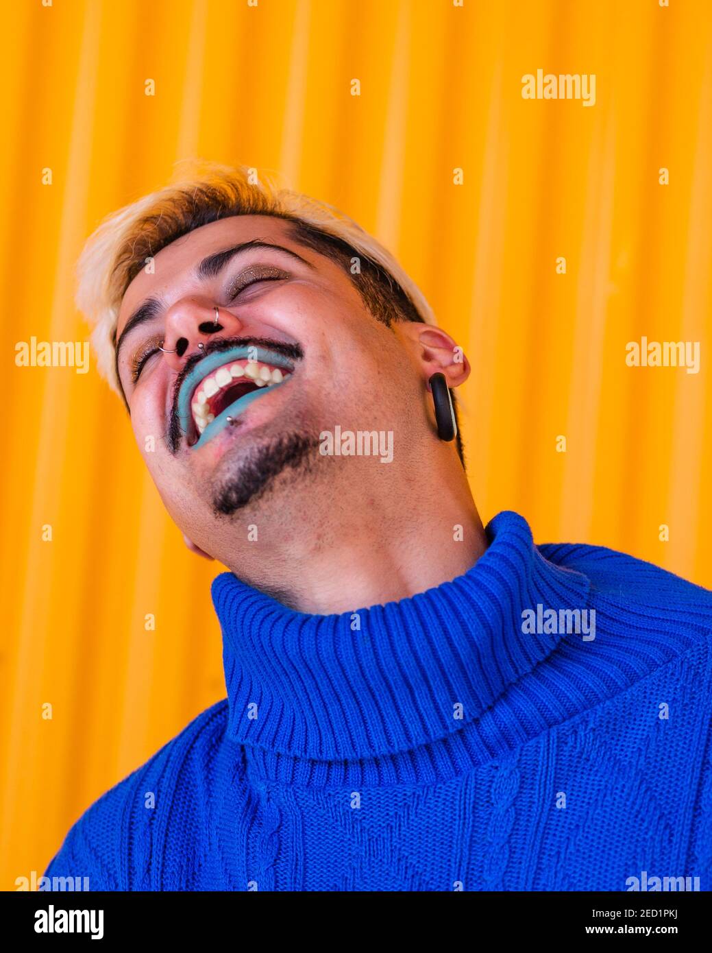 Androgynous cheerful male with blue lips and in stylish wear standing on background of yellow wall in city with eyes closed Stock Photo