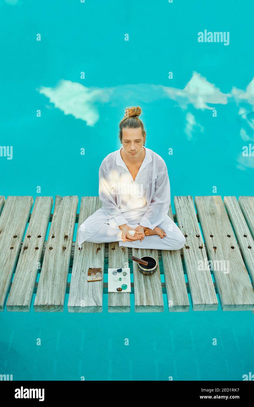 From above blond male hipster yogi in white clothes sitting in lotus pose meditating near tibetan singing bowl and crystals on wooden path bridge on t Stock Photo