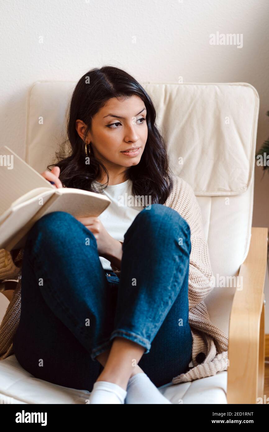 Female musician sitting in armchair and writing in notebook while composing music and looking away in contemplation Stock Photo