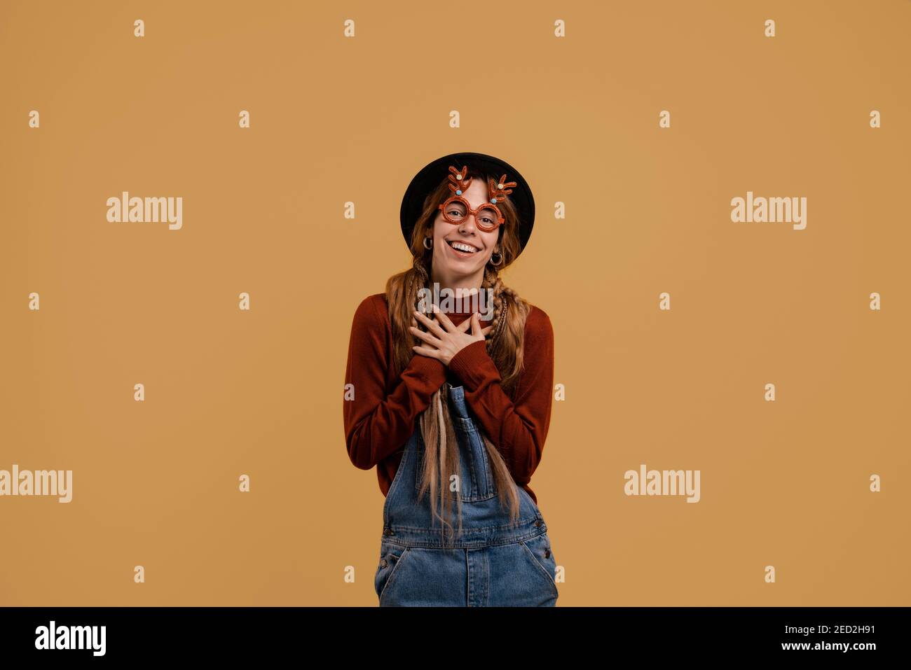 Photo of attractive female farmer looks pleasant and happy, wears funny glasses. White woman wears denim overall and hat isolated brown color Stock Photo