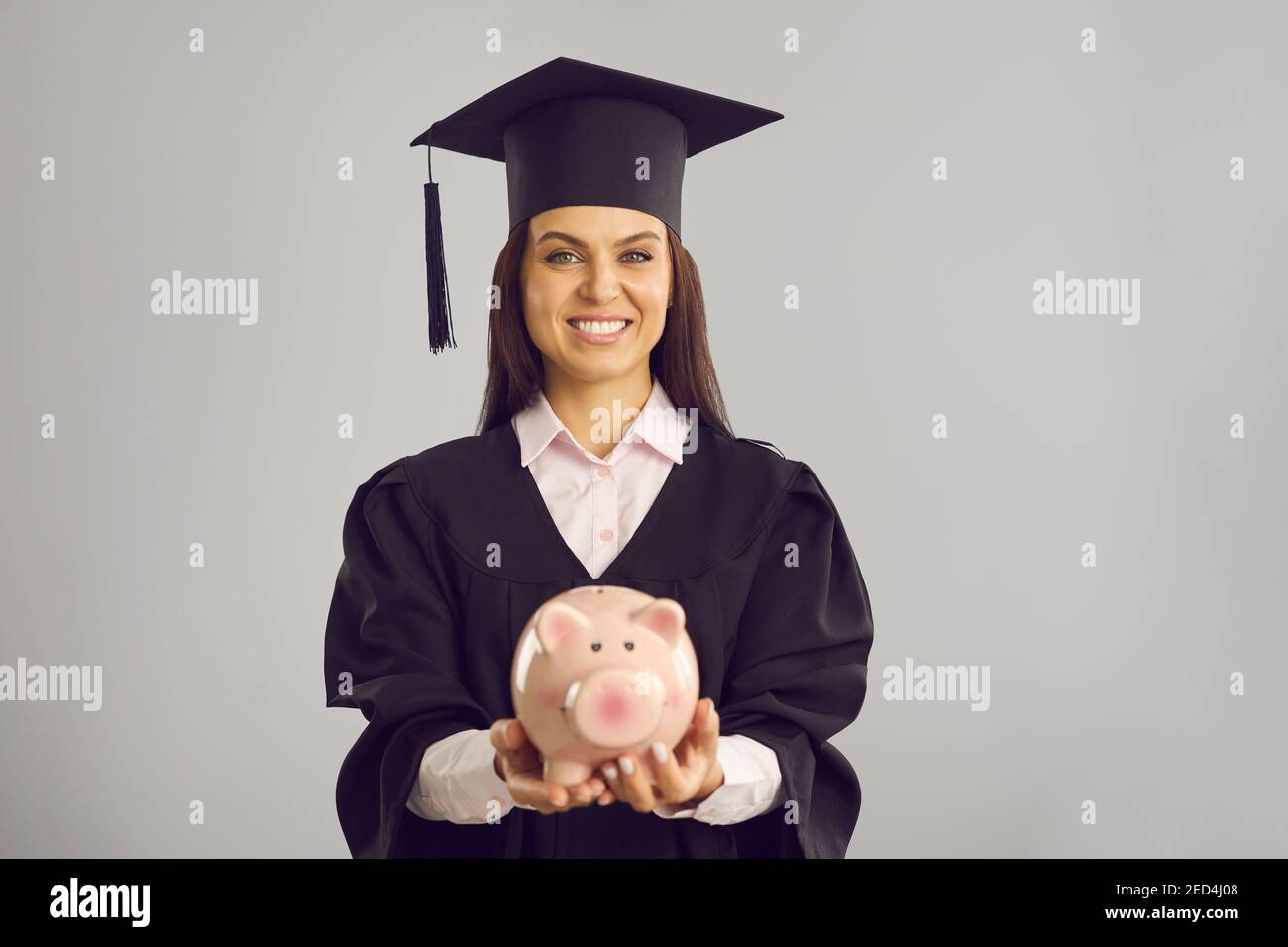 Happy student in academic cap and gown holding piggy bank with money saved for education Stock Photo