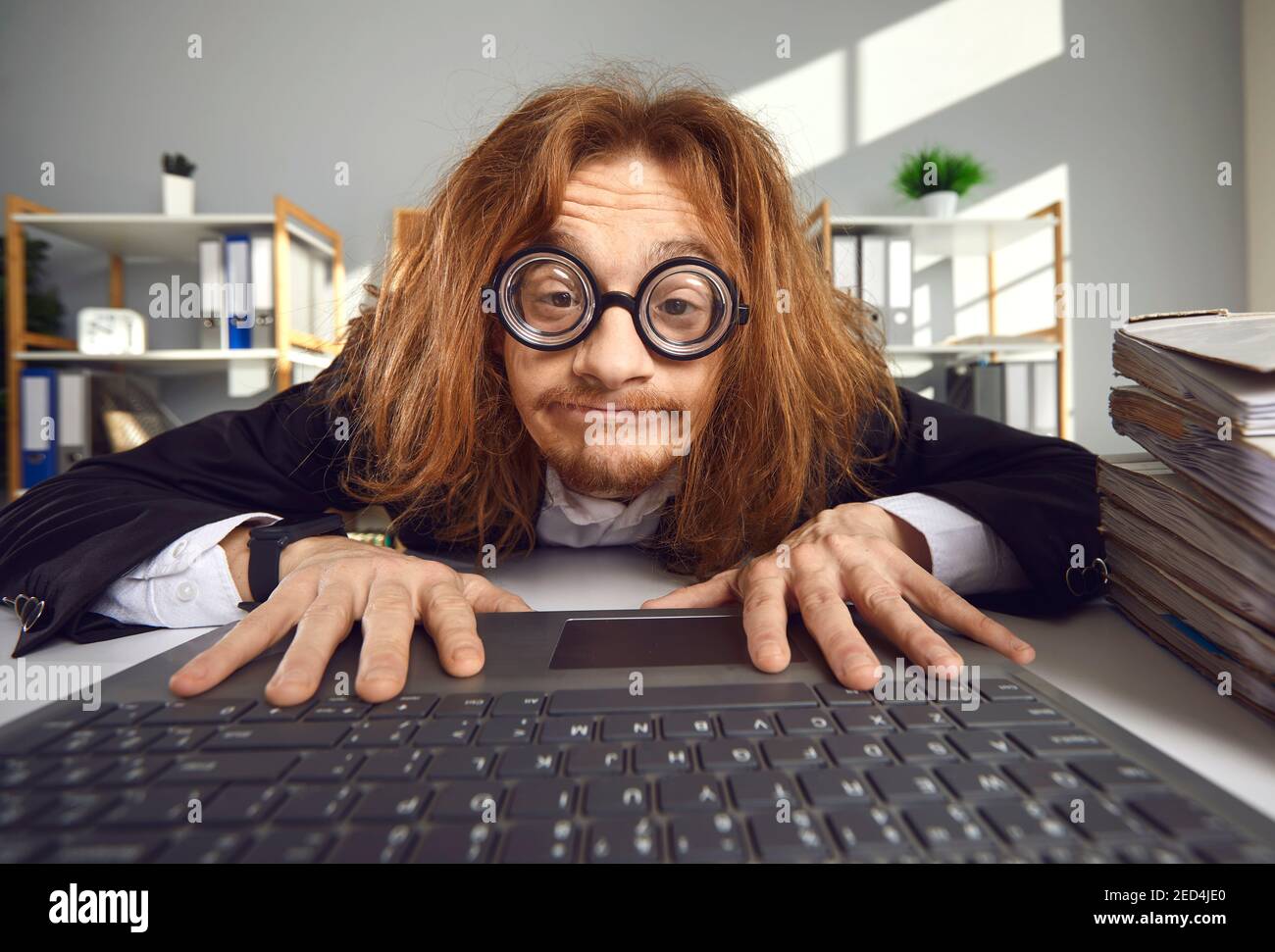 Funny crazy looking nerd in thick lens glasses sitting in office and using laptop Stock Photo